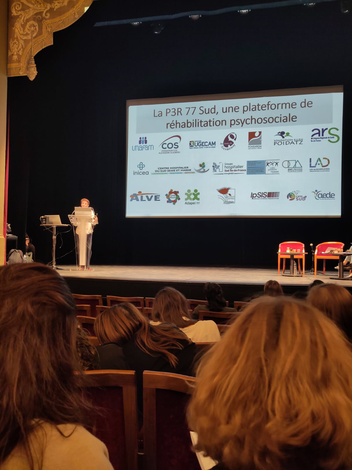 Conférence dans un théâtre : une femme présente sur scène devant un public, avec une diapositive affichant des logos et le titre d’une plateforme de réhabilitation psychosociale.
