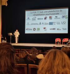 Conférence dans un théâtre : une femme présente sur scène devant un public, avec une diapositive affichant des logos et le titre d’une plateforme de réhabilitation psychosociale.
